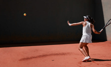 Female tennis player in white outfit serving on red clay court, focused on ball mid-air